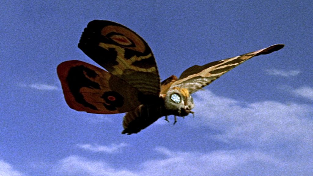 This is a film still from MOTHRA モスラ dir Ishirō Honda (1961) showing a giant colourful moth flying across a blue sky.