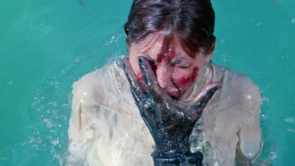 This is a film still from STING OF DEATH dir William Grefé (1966), showing a fully-clothed woman emerging from a pool of water with a bloodied face covered by the hand of an unseen monster.