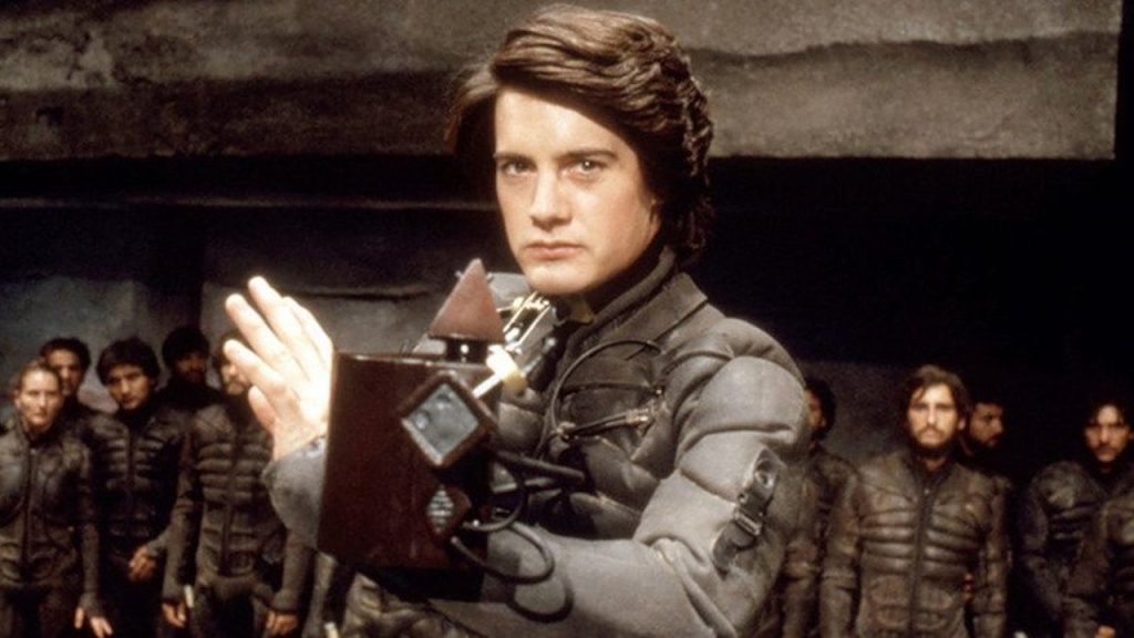 This is a film still from David Lynch's DUNE (1984) showing Kyle MacLachlan as Paul Atreides, in his battle stance, wearing a stillsuit and holding a weirding module, a group of Fremen stand in a row behind him, looking on in awe.