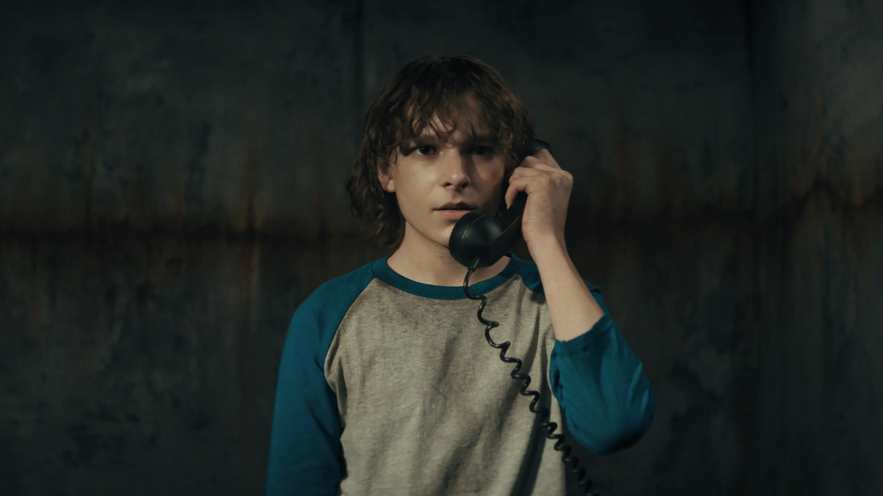This is a film still from THE BLACK PHONE (2021) showing Mason Thames as Finney holding a phone to his ear as he stands alone in a basement.