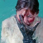 This is a film still from STING OF DEATH dir William Grefé (1966), showing a fully-clothed woman emerging from a pool of water with a bloodied face covered by the hand of an unseen monster.