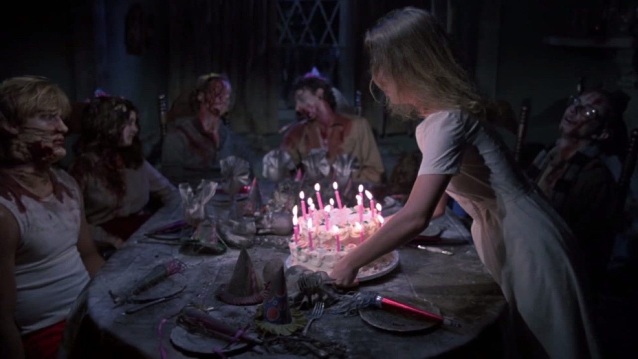 This is a film still from HAPPY BIRTHDAY TO ME (1981) showing a birthday cake with candles lit being placed on a table surrounded by seated corpses.