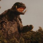 This is a film still from GAMERA, GUARDIAN OF THE UNIVERSE showing a giant fire-breathing turtle standing on its hind legs as it emerges from a forest.