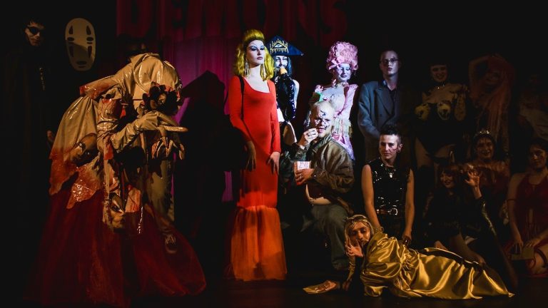 This is a photograph from the costume parade at the Queer Horror Nights screening of DEMONS for Halloween, showing a number of audience members posing together in costume on the main stage at Rio Cinema, looking into the auditorium and waiting to be judged!