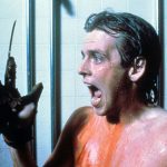 This is a film still from A NIGHTMARE ON ELM STREET 2 - FREDDY'S REVENGE dir Jack Sholder (1985) showing a teenage boy standing in the school showers, covered in blood. He's holding up his hand on which he's wearing Freddy Kreuger's knifed glove, and he's screaming in terror.