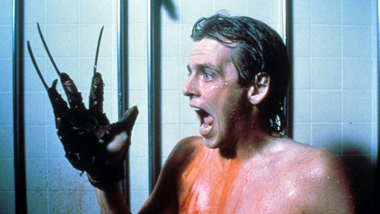 This is a film still from A NIGHTMARE ON ELM STREET 2 - FREDDY'S REVENGE dir Jack Sholder (1985) showing a teenage boy standing in the school showers, covered in blood. He's holding up his hand on which he's wearing Freddy Kreuger's knifed glove, and he's screaming in terror.