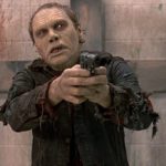 This is a film still from George A Romero's DAY OF THE DEAD, showing a zombie holding a revolver and pointing it threateningly at someone directly in front of them.