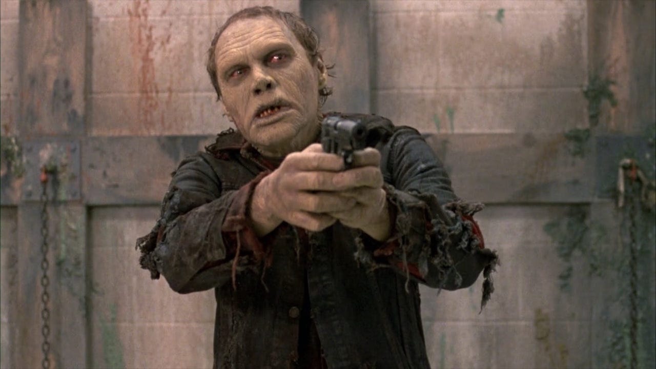 This is a film still from George A Romero's DAY OF THE DEAD, showing a zombie holding a revolver and pointing it threateningly at someone directly in front of them.