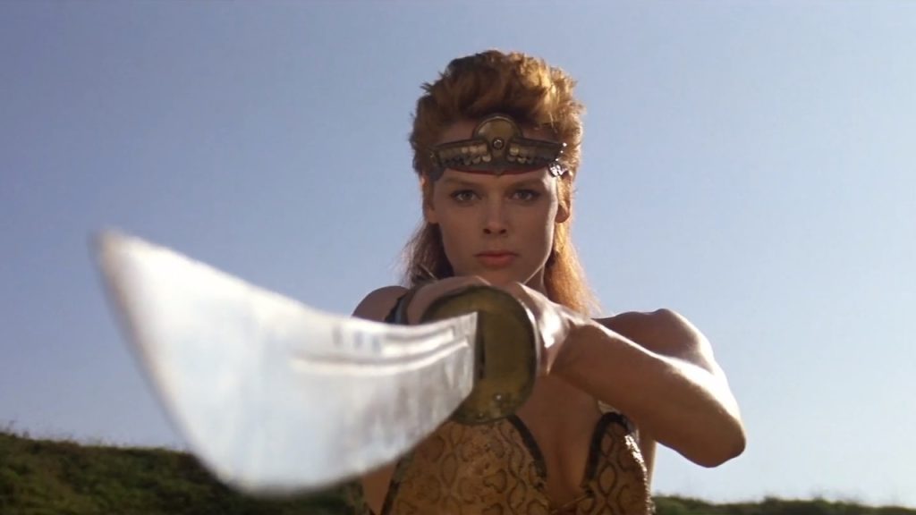 This is a film still from RED SONJA (1985) showing the actress Brigitte Nielsen in character as Red Sonja standing in a warrior pose, sword pointing to the camera.
