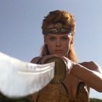 This is a film still from RED SONJA (1985) showing the actress Brigitte Nielsen in character as Red Sonja standing in a warrior pose, sword pointing to the camera.