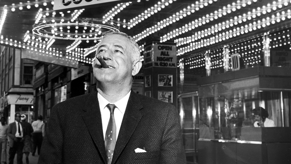 This is a black and white photograph of film director William Castle, showing the film gimmick pioneer standing in a suit and tie while smoking a cigar in front of a 1960s cinema marquee all lit up for the night.