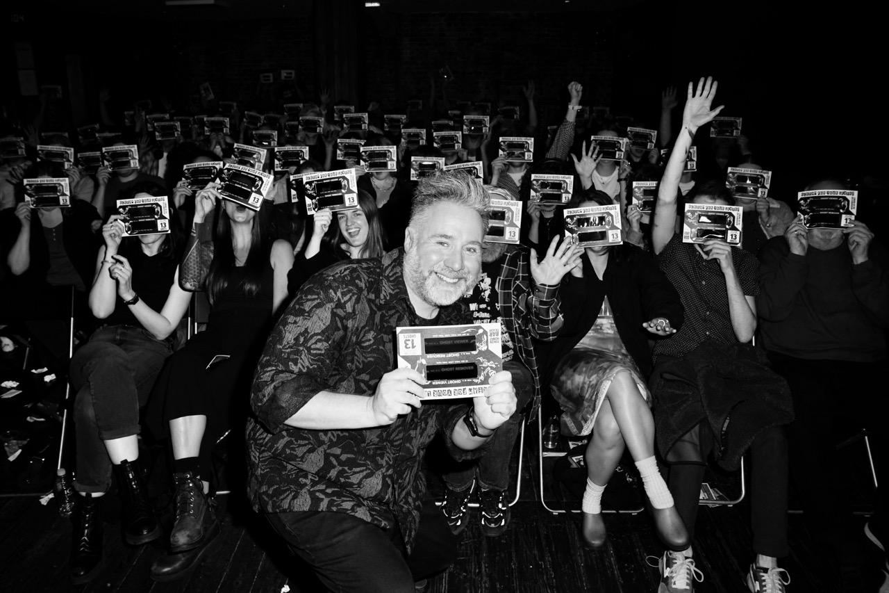 This is a picture of Bar Trash host Token Homo holding up a cardboard 'ghost viewer' and smiling at the camera whilst standing in front of the Bar Trash audience all holding their viewers to thier faces ready to watch William Castle's film, 13 GHOSTS.