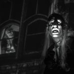This is a black and white photograph of London-based drag creature Co Kendrah performing in costume as Riff-Raff in front of a screening of THE ROCKY HORROR PICTURE SHOW at Rio Cinema in Dalston. We can see Richard O'Brien performing on the screen behind them during the song 'Over at the Frankenstein Place'.